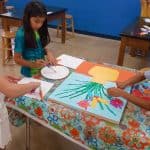 Title: Mary Jo’s Flower Farm
Size: 18” x 24”
Medium: acrylic paint, acetate paper
Description:  Inspired by their field trip with kindergarten science buddies to Mary Jo’s Flower Farm, each second grade artist designed a colorful flower using acetate paper. Students worked in small groups to paint a foreground, background, and vase. After painting individual stems, students attached their flowers to the composition. As the final step in their creative process, the class brainstormed title ideas for their piece then voted for their favorite.
