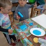 Title: The Colorful Ghosts
Size: 24”x18”
Media: acrylic paint
Description: 1st grade artists used printmaking to create a colorful quilt-like composition. Each student rolled paint onto a rectangular piece of foam and stamped the shape onto the wooden panel. After designing a printmaking block with foam shapes, they rolled paint onto it and stamped it directly over their solid block of color. Students repeated this process with a partner to create a second print. These student pairs practiced sharing their ideas for design and color choices. The last printed block was created by the entire class. As the final step in their creative process, the class brainstormed title ideas for their piece then voted for their favorite.
