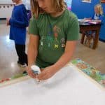 Title: Shapes Collide
Size: 24”x18”
Medium: fabric, acrylic paint
Description: 5th grade students began their batik creation by drawing a simple line and shape design on a small rectangle of paper and tracing it with black Sharpie. After the papers were taped onto a piece of cardboard, covered with wax paper, and finally covered with a large piece of white fabric, students carefully traced their designs with glue onto the fabric. Once the glue was dry, students painted the sections of their design with acrylic paint. When the paint was dry, the glue was washed out of the fabric to reveal the white outline left by the glue. As the final step in their creative process, the class brainstormed title ideas for their piece then voted for their favorite.
