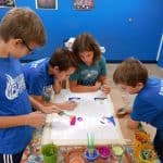 Title: Shapes Collide
Size: 24”x18”
Medium: fabric, acrylic paint
Description: 5th grade students began their batik creation by drawing a simple line and shape design on a small rectangle of paper and tracing it with black Sharpie. After the papers were taped onto a piece of cardboard, covered with wax paper, and finally covered with a large piece of white fabric, students carefully traced their designs with glue onto the fabric. Once the glue was dry, students painted the sections of their design with acrylic paint. When the paint was dry, the glue was washed out of the fabric to reveal the white outline left by the glue. As the final step in their creative process, the class brainstormed title ideas for their piece then voted for their favorite.
