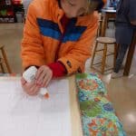 Title: Shapes Collide
Size: 24”x18”
Medium: fabric, acrylic paint
Description: 5th grade students began their batik creation by drawing a simple line and shape design on a small rectangle of paper and tracing it with black Sharpie. After the papers were taped onto a piece of cardboard, covered with wax paper, and finally covered with a large piece of white fabric, students carefully traced their designs with glue onto the fabric. Once the glue was dry, students painted the sections of their design with acrylic paint. When the paint was dry, the glue was washed out of the fabric to reveal the white outline left by the glue. As the final step in their creative process, the class brainstormed title ideas for their piece then voted for their favorite.
