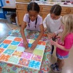 Title: The Colorful Ghosts
Size: 24”x18”
Media: acrylic paint
Description: 1st grade artists used printmaking to create a colorful quilt-like composition. Each student rolled paint onto a rectangular piece of foam and stamped the shape onto the wooden panel. After designing a printmaking block with foam shapes, they rolled paint onto it and stamped it directly over their solid block of color. Students repeated this process with a partner to create a second print. These student pairs practiced sharing their ideas for design and color choices. The last printed block was created by the entire class. As the final step in their creative process, the class brainstormed title ideas for their piece then voted for their favorite.
